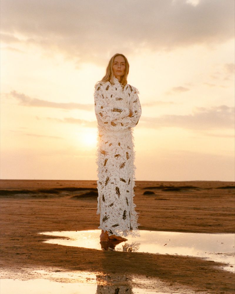 Model Georgina Grenville standing barefoot in a long textured white coat on a sandy shore at sunset.