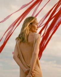 Model Georgina Grenville in a backless beige dress standing outdoors beneath red streamers blowing across the sky.
