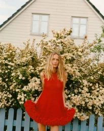 Model Georgina Grenville poses in a red knit ruffle-hem mini dress in front of flowering bushes and a blue fence.