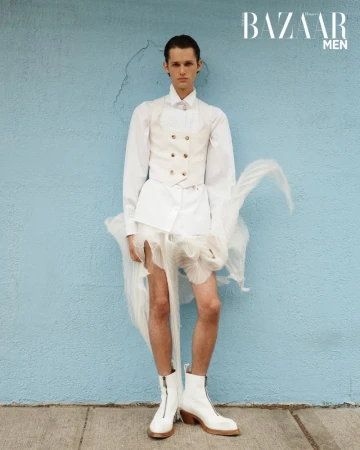 Charlie Nolte posing in a white layered vest, sheer fabric skirt, and white boots against a light blue wall.