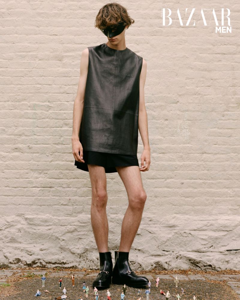Man in black leather top and eye mask stands in front of brick wall with miniature people on ground in a fashion editorial shot.