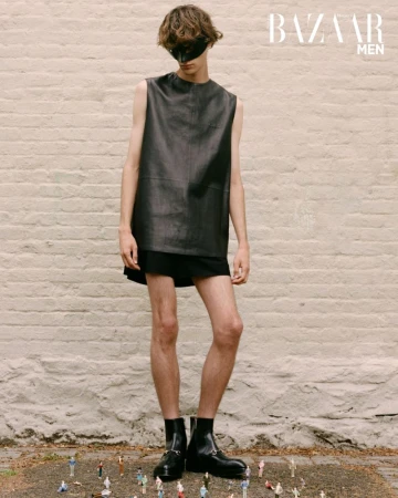 Man in black leather top and eye mask stands in front of brick wall with miniature people on ground in a fashion editorial shot.