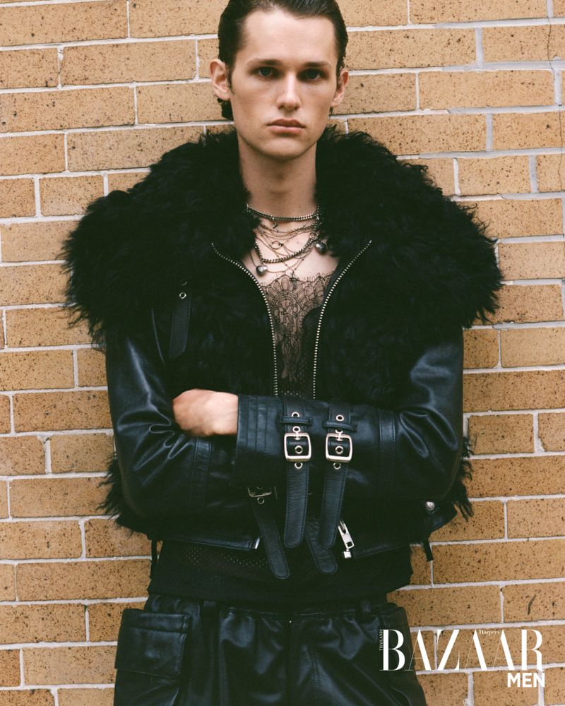 Fashion portrait of a male model in a black leather jacket with a fur collar posing against a brick wall for a magazine cover.