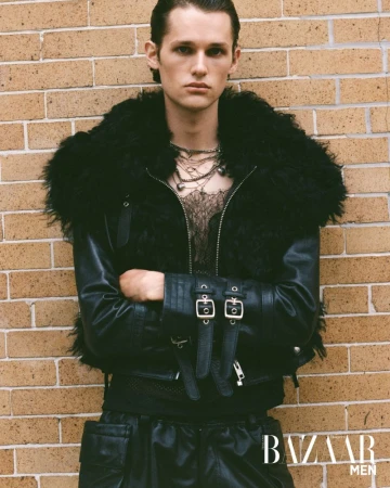 Fashion portrait of a male model in a black leather jacket with a fur collar posing against a brick wall for a magazine cover.