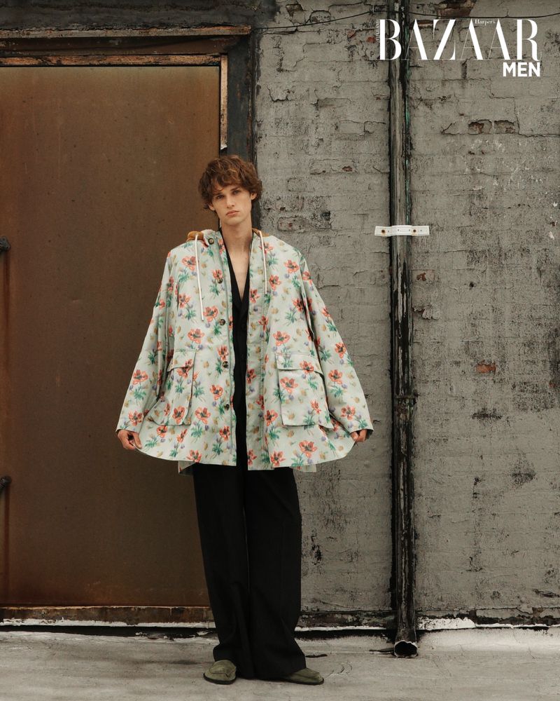 A young male model poses outdoors in a floral hooded jacket, black pants, and green shoes, embodying a casual yet fashionable look.