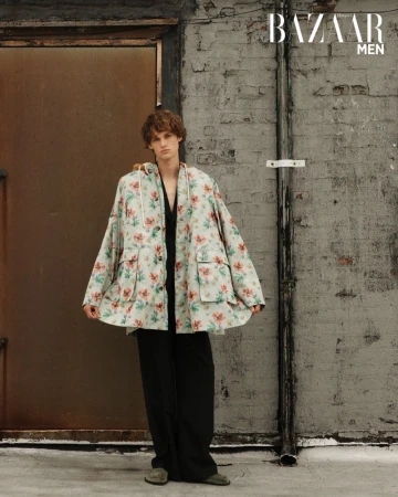 A young male model poses outdoors in a floral hooded jacket, black pants, and green shoes, embodying a casual yet fashionable look.