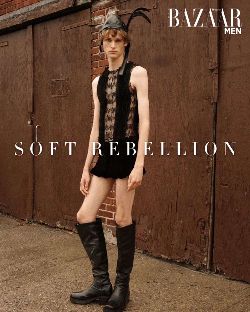 Fashion editorial shot of a slender male model in a lace top, black shorts, leather headdress, and black boots, posing outdoors against a brown brick wall.
