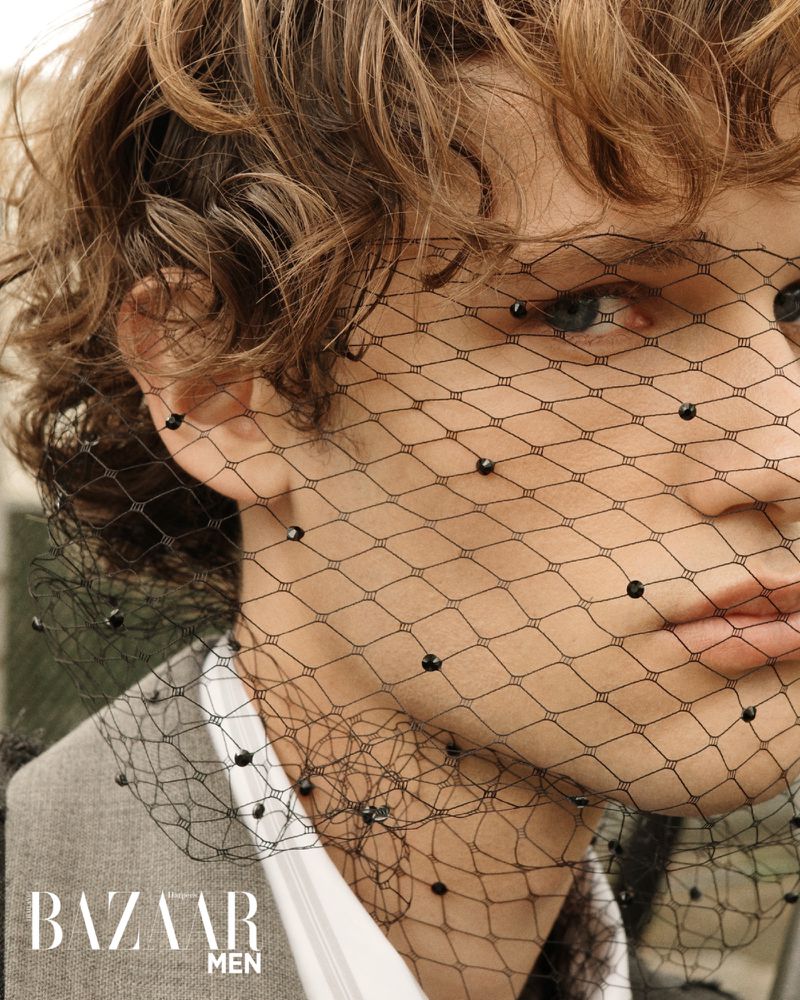 Close-up of a person wearing a beaded net over their face, with curly hair, a grey suit, and the Bazaar Men logo visible.