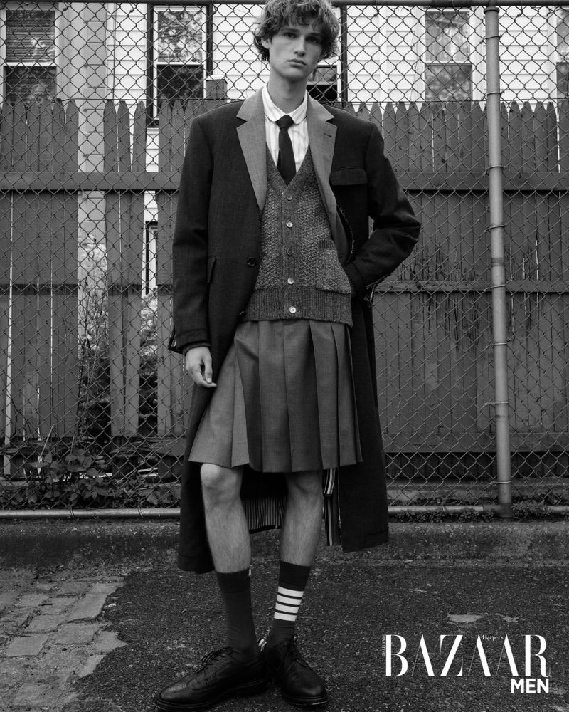 Black and white portrait of a young man modeling outdoors in a coat, vest, tie, shirt, and kilt with a chain link fence backdrop.