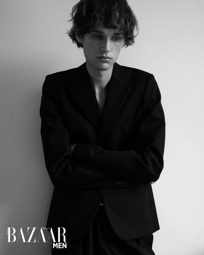 Black and white portrait of a young adult man in a black suit leaning against a wall. Bazaar Men logo visible.