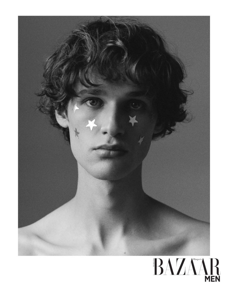 Black and white headshot of a young adult male with curly hair and star-shaped stickers on his face against a grey background.