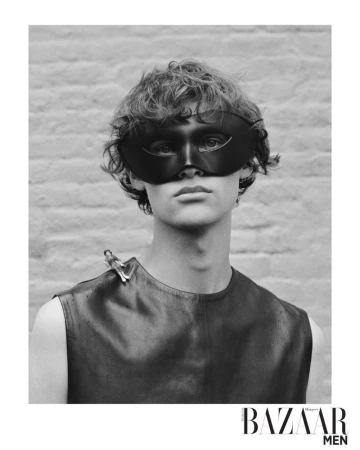 Monochrome portrait of a male fashion model wearing a black leather mask and top in front of a light brick wall for Bazaar Men.