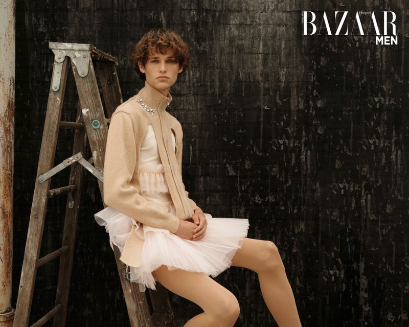 Young adult male model poses in a ballet tutu with a ladder in the background wearing a nude cardigan for Bazaar Men.