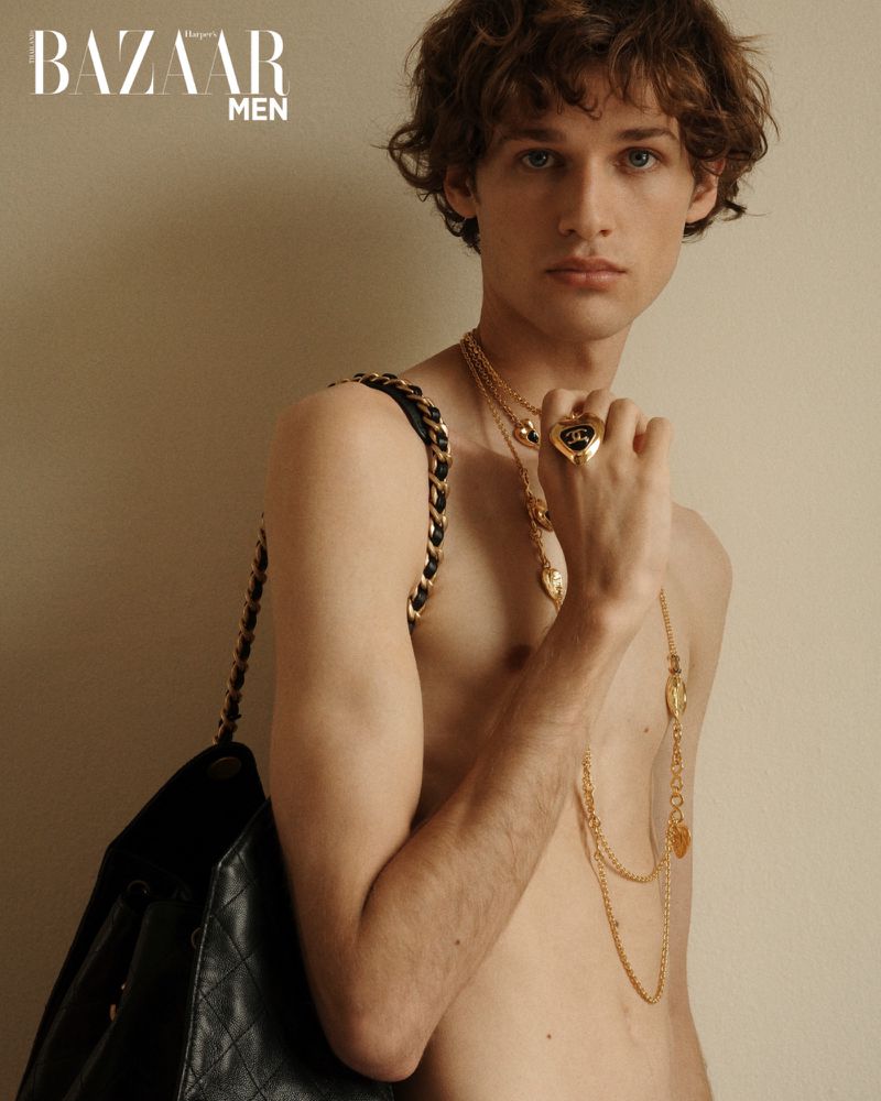 Portrait of a slender male fashion model with curly brown hair, wearing gold jewelry and carrying a quilted backpack, against a neutral background.
