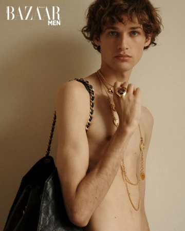 Portrait of a slender male fashion model with curly brown hair, wearing gold jewelry and carrying a quilted backpack, against a neutral background.