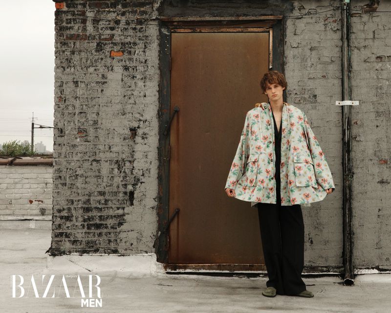 Editorial fashion shot of a young man in a floral print coat standing on a rooftop against a brick wall with a metal door.