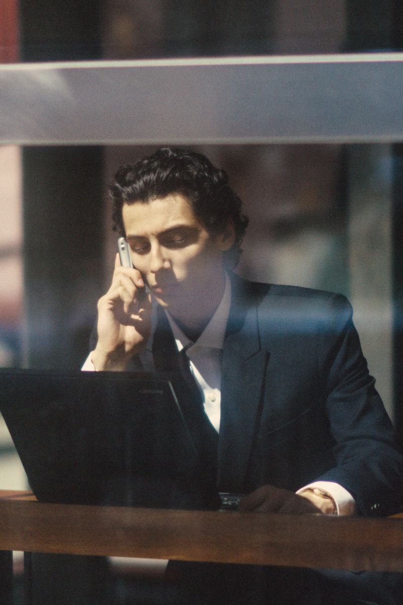 Man in a dark suit talking on a cell phone while sitting at a desk with a laptop, seen through a reflective window.