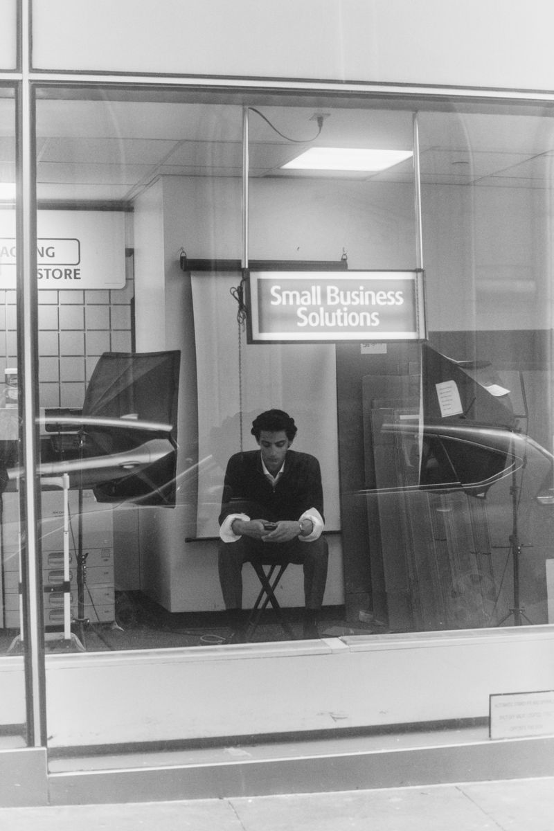 Mehdi Abouzaid seated inside a storefront beneath an illuminated Small Business Solutions sign, photographed in black and white.