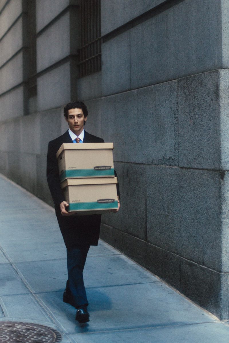 Man in a dark tailored overcoat and tie walking on a city sidewalk while carrying two stacked cardboard storage boxes.