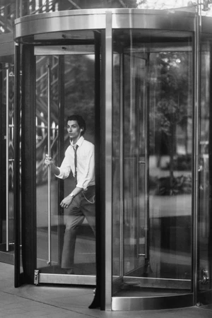 Black and white photo of a man in a shirt and tie walking through a glass revolving door while wearing earphones.