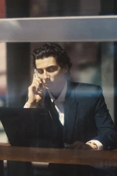 Man in a dark suit talking on a cell phone while sitting at a desk with a laptop, seen through a reflective window.