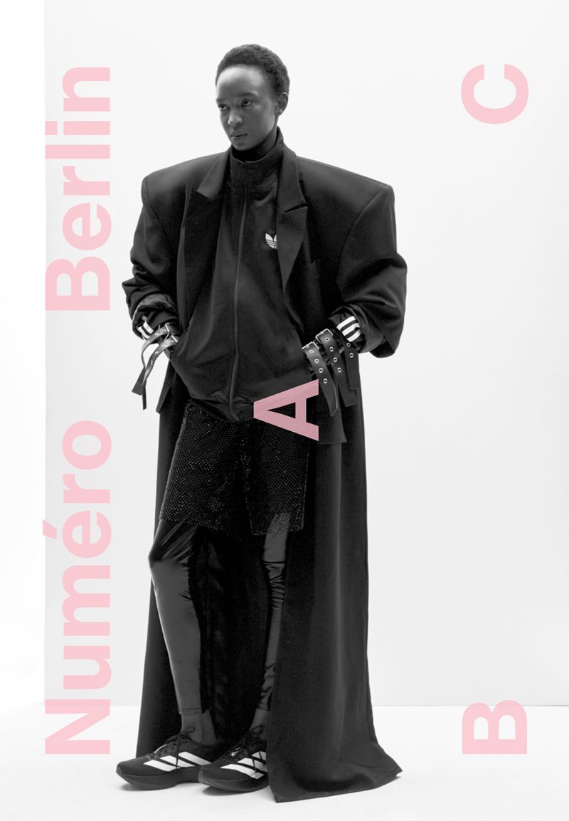 Editorial fashion shot of a Black female model in a full-length black Adidas tracksuit and long black coat, posed in a white studio.