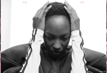 Black and white portrait of model Nicole Atieno with hands on her head wearing a high-shoulder jacket against a white background.