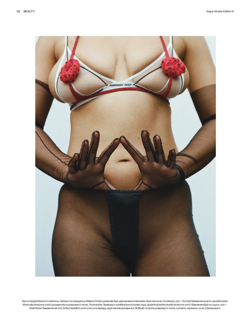 Close-up of a model wearing a sheer white bra with red rosette appliqués and sheer black gloves against a pale studio backdrop.