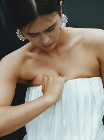 Model Noor Mutsaers looking down wearing a strapless white pleated dress and crystal cluster earrings against a dark backdrop.