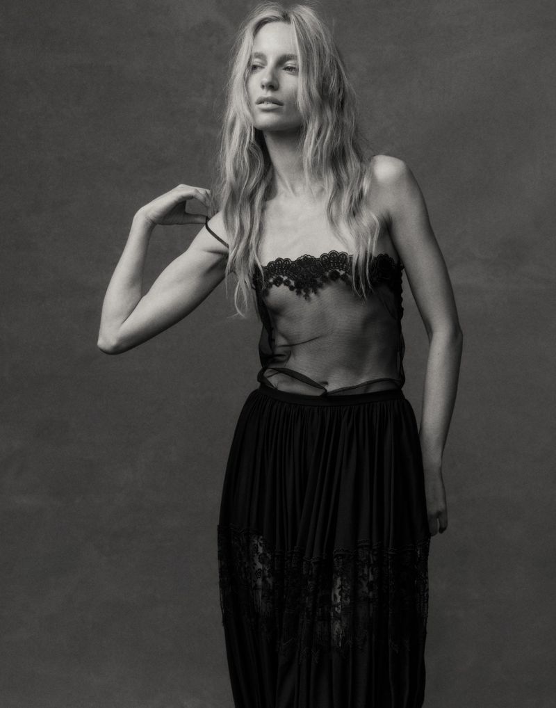 Monochrome fashion portrait of a tall, slender woman in a sheer lace top and long skirt, posing against a textured grey background.