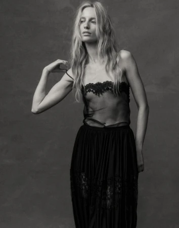Monochrome fashion portrait of a tall, slender woman in a sheer lace top and long skirt, posing against a textured grey background.