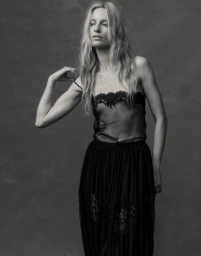 Black and white portrait of Maya Stepper wearing a sheer black lace top and dark skirt against a textured studio backdrop.