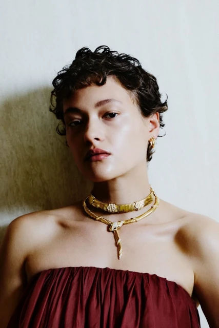 A woman with curly hair wearing a strapless top and a gold snake necklace poses against a neutral background in a medium shot.