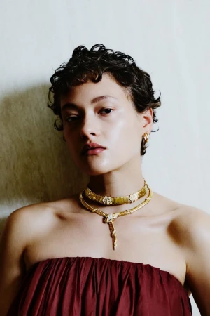 Daria Zolotova wearing a gold Bulgari Serpenti necklace and strapless burgundy dress against a neutral textured studio backdrop.