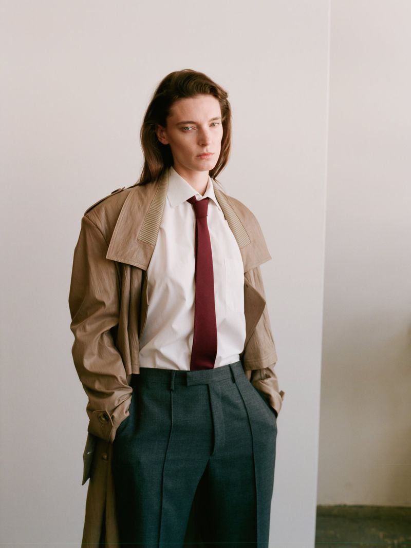 Model Ansley Gulielmi posing in a tan trench coat, white collared shirt, burgundy tie, and tailored dark trousers.