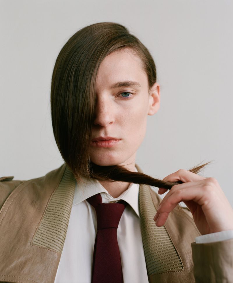 Model Ansley Gulielmi holding hair across face, wearing a tan jacket, white collared shirt, and burgundy tie against a pale backdrop.