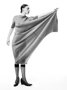 Model Ansley Gulielmi poses in an extended sculptural pleated grey dress and black heels against a white studio backdrop.