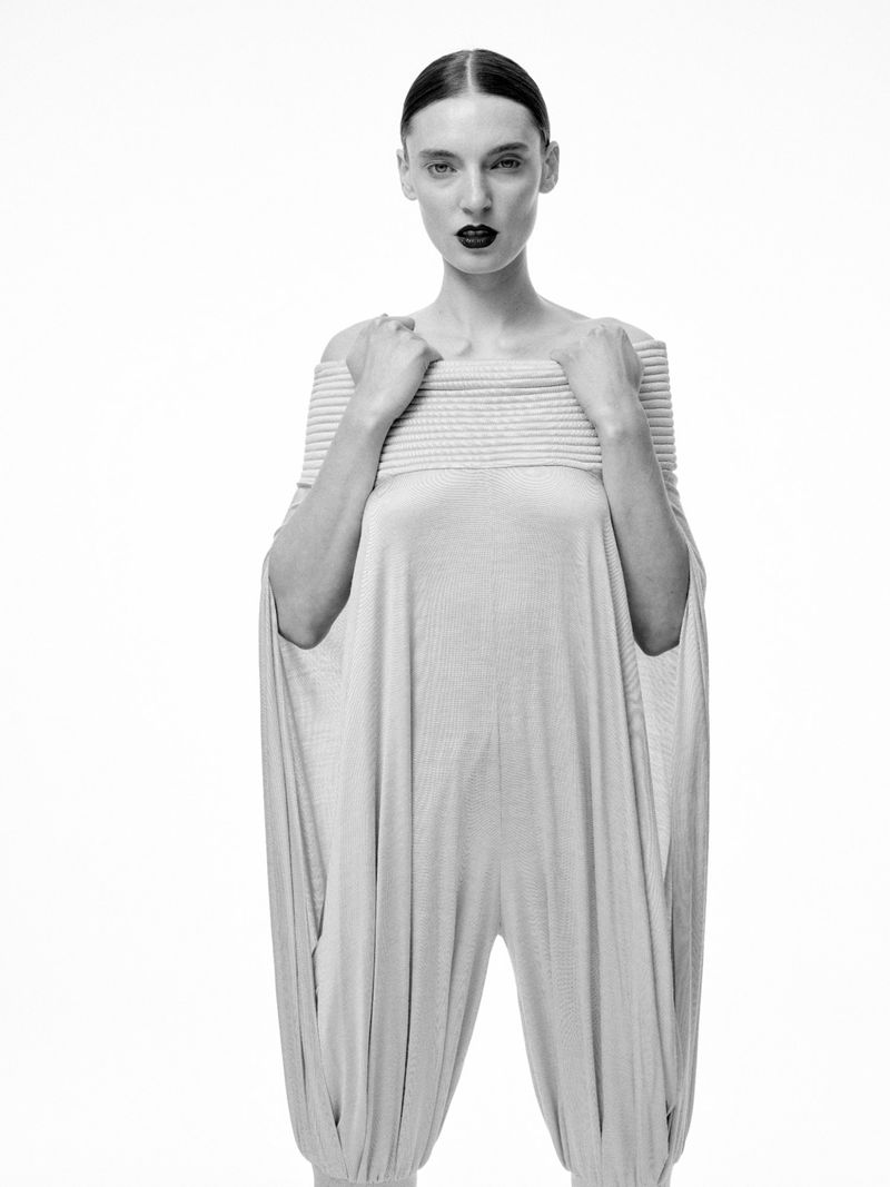Model Ansley Gulielmi wearing a light grey off-the-shoulder draped jumpsuit with dark lipstick against a white studio background.
