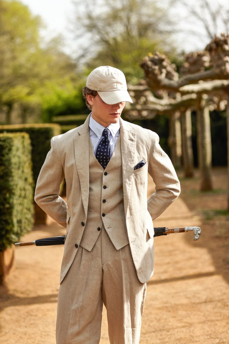Adult male in linen suit and cap stands in a garden with an umbrella cane, capturing a blend of formal wear and outdoor lifestyle.