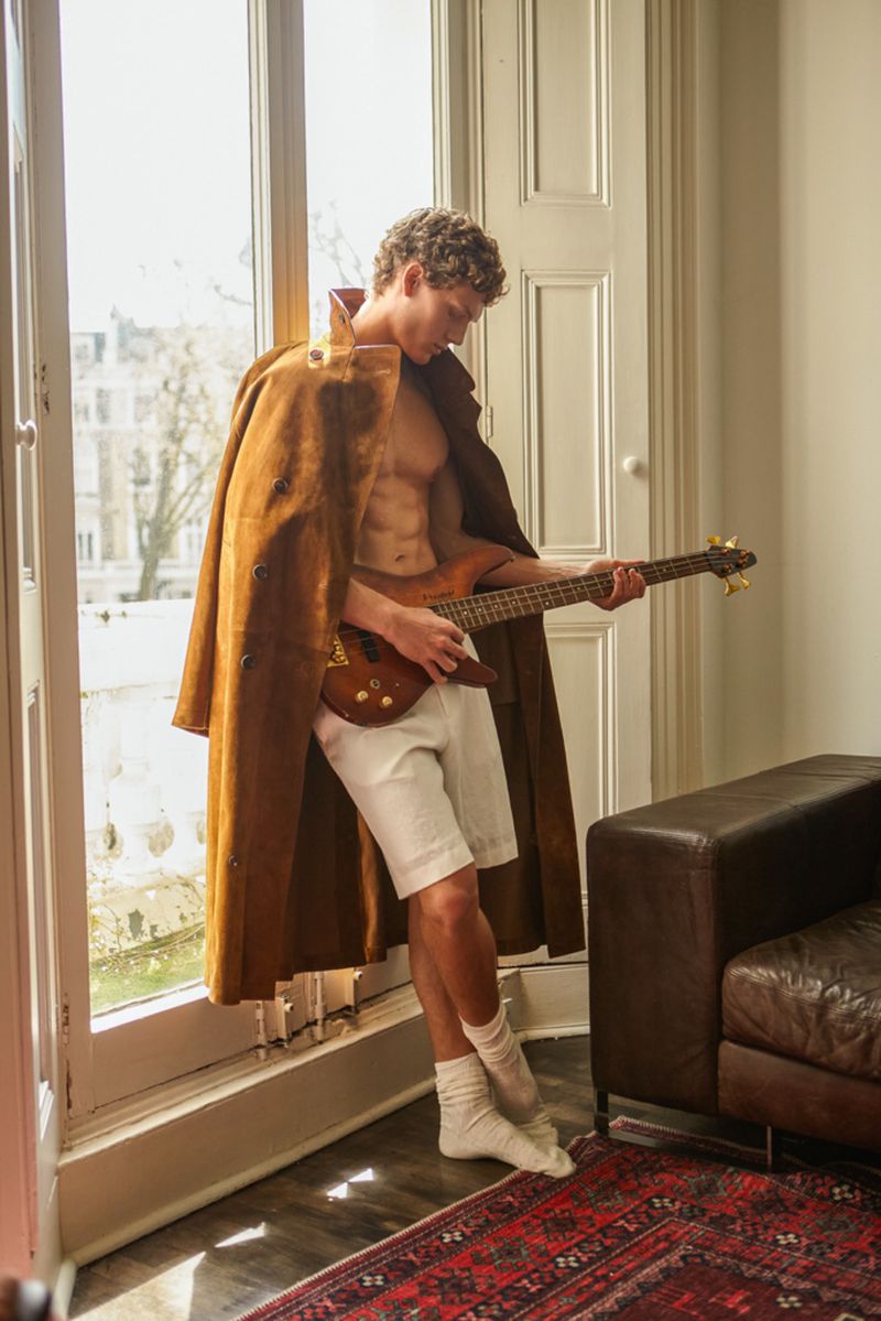 An adult male plays an electric bass guitar next to a window while wearing a brown coat, white shorts, and socks inside a room.