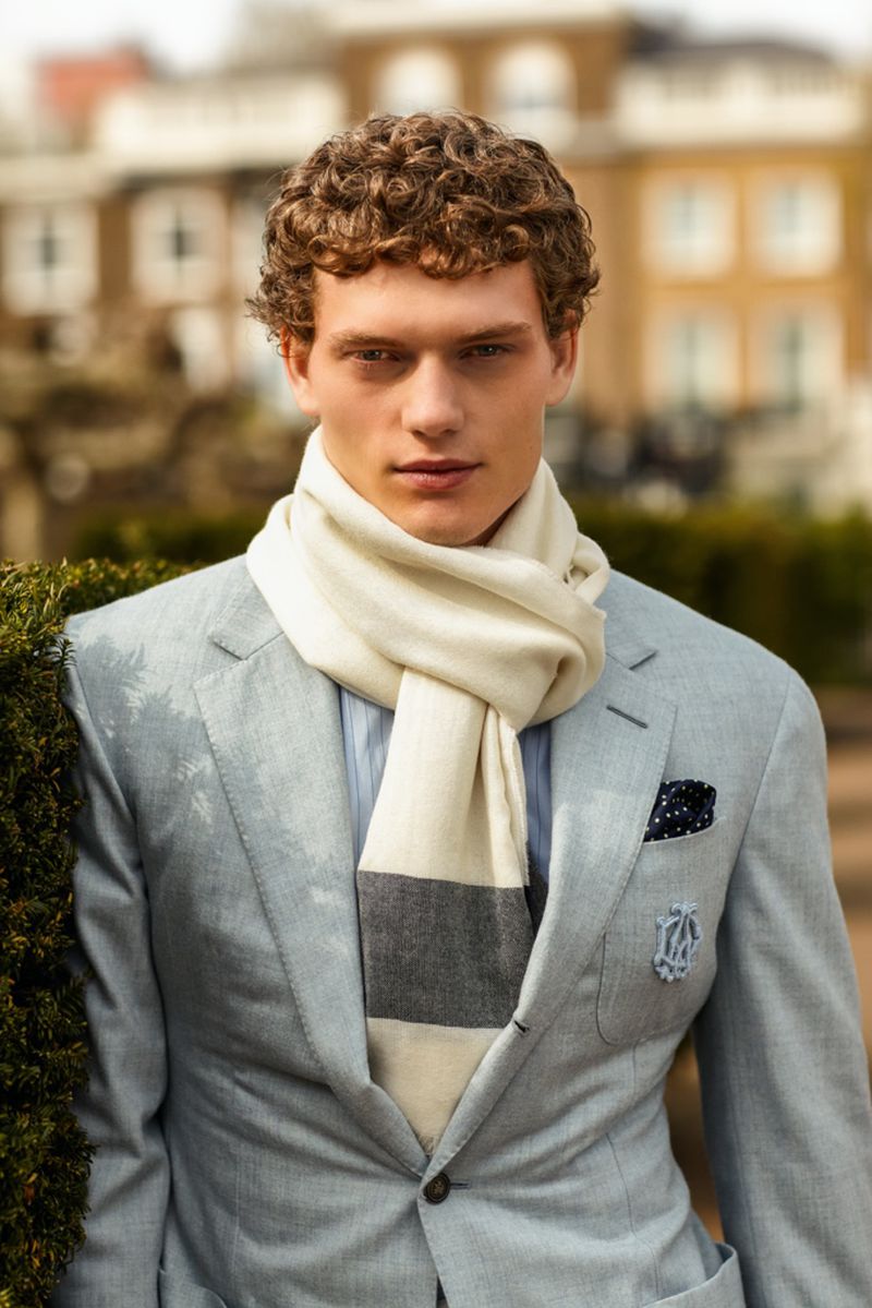 Male fashion model wearing a light blue suit and cream-colored scarf poses outdoors, featuring a blurred building backdrop.