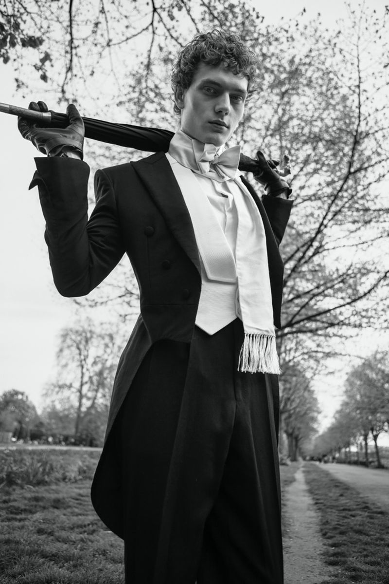 Black and white portrait of an adult man in a vintage-style formal tuxedo and holding an umbrella outdoors on a path.