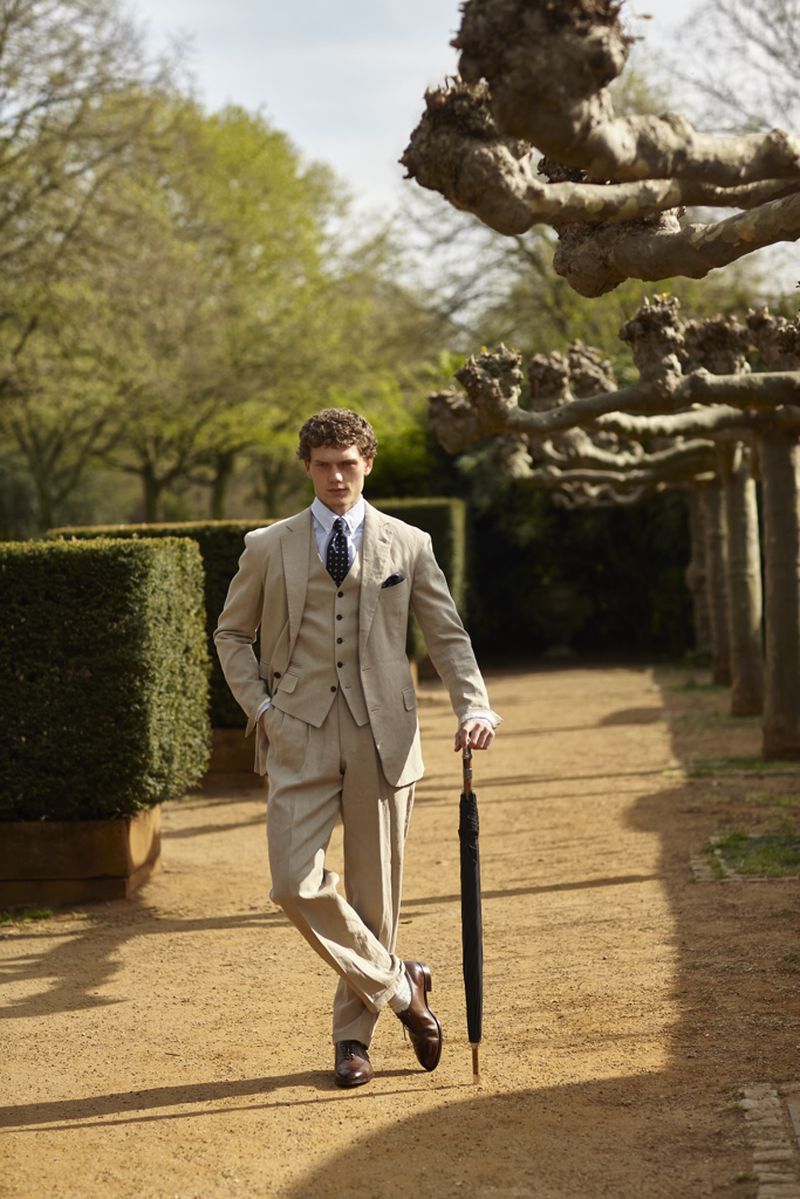 Man in a beige suit stands on a path in a park, holding an umbrella. Well-groomed hedges and bare trees line the walkway on a sunny day.