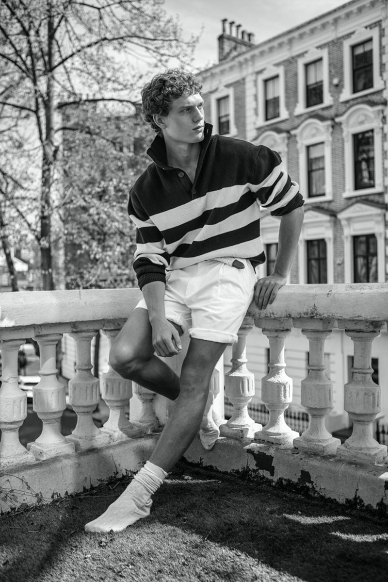 Black-and-white shot of a curly-haired adult male leaning on a balustrade with buildings and trees in the background looking away.