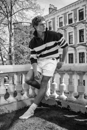 Black-and-white shot of a curly-haired adult male leaning on a balustrade with buildings and trees in the background looking away.