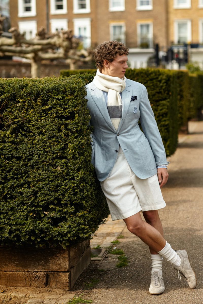 Male model in a light blue blazer and white shorts leans against a green hedge in an outdoor setting with buildings in the background.