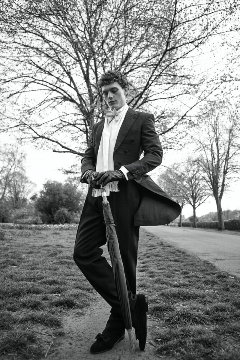 Monochrome portrait of a man in a black suit and bow tie holding an umbrella outdoors with trees and a path in the background.