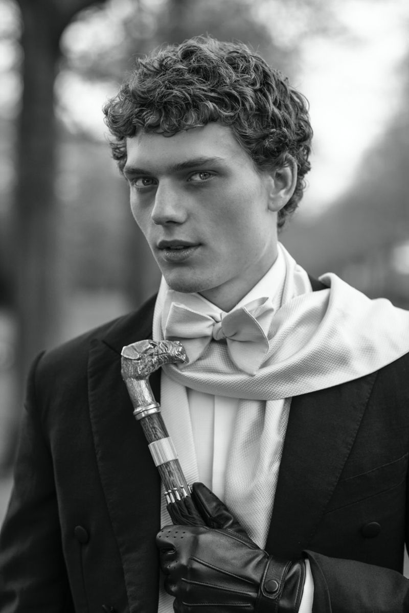 Monochrome portrait of a stylish adult male in a tuxedo with a bow tie, holding a walking cane, outdoors.