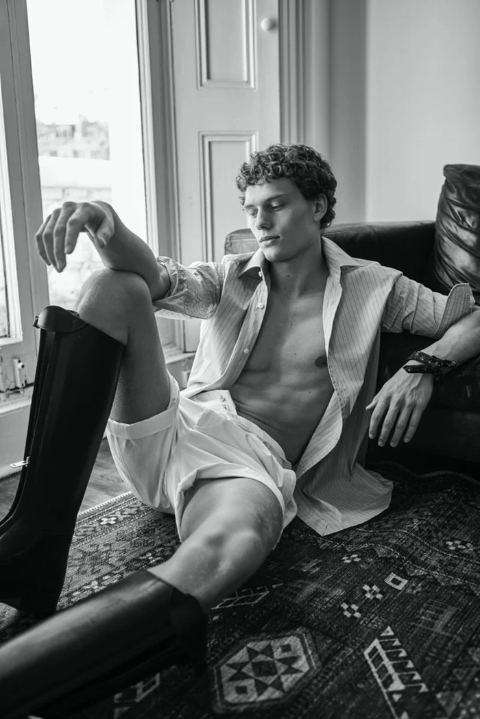 A young male model seated on a patterned rug wearing an unbuttoned striped shirt and tall boots, photographed for Dunhill.