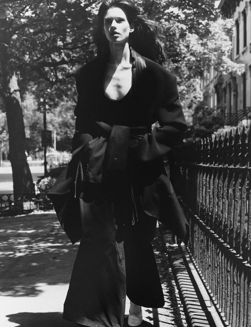 Model Sandra Murray walking outdoors in a black draped tailored coat beside a black iron fence in black and white.